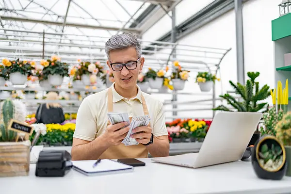 Çiçekçi dükkanındaki beyaz bir adam kazandıklarını sayıyor. Etrafı renkli çiçekler ve satılık bitkilerle çevrili. Tezgahta..