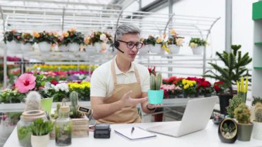 Videodaki bir çiçekçi, müşteriye kaktüsü gösteriyor. Dükkanda bir sürü çiçek var..