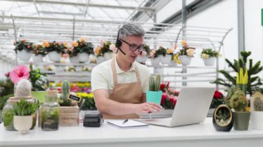 Kafkasyalı bir çiçekçi, satılık bir kaktüsü sergilerken bir video görüşmesi yapıyor ve bir dizüstü bilgisayarla online satışları vurguluyor..