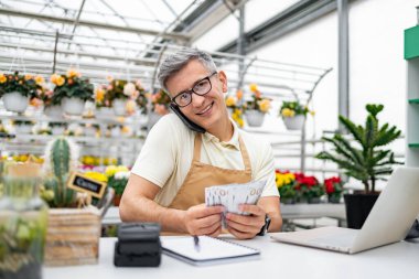 Kafkasyalı bir çiçekçi, elinde parayla mutlu, dükkanında çiçeklerle çevrili bir telefon görüşmesi yaparken gülümsüyor..