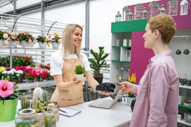Çiçekçi, bir çiçekçinin kasasında müşteriye yardım ediyor. Gülümseyen bir çalışan küçük bir saksı bitkisinin yanından geçiyor..