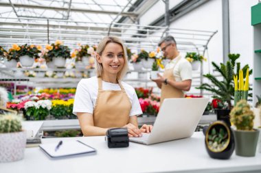 Bir çiçekçi işte, gülümsüyor ve dizüstü bilgisayar kullanıyor. Arka planda, bir meslektaşım çiçeklerle dolu güneşli bir serada bir bitkiyi inceliyor..