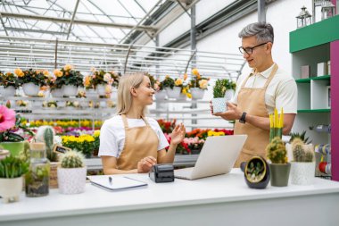 Beyaz bir erkek ve kadın çiçekçide, küçük bir bitkiye bakarken birbirleriyle etkileşim halindedirler..