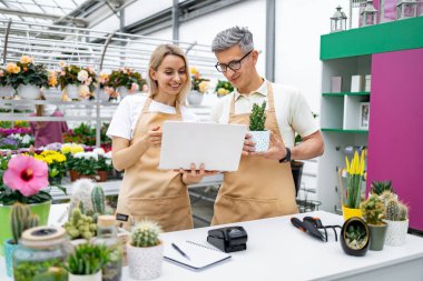 Bir çiçekçi ve bir müşteri mağazadaki satıştayken dizüstü bilgisayarı kontrol ediyorlar. Kadın dizüstü bilgisayarı tutuyor, ve adam saksı bitkisini tutuyor..