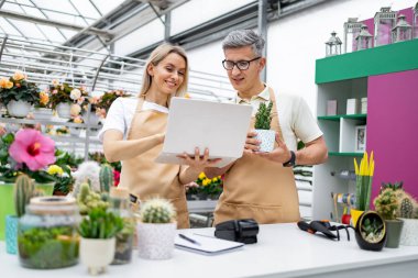 İki çiçekçi, bir erkek ve bir kadın, bir fabrikada dizüstü bilgisayara bakıyorlar, masada saksıdaki küçük bir bitkiden bahsediyorlar..