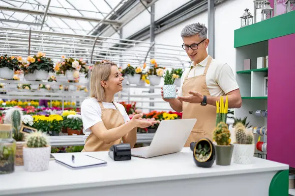 İki çiçekçi, çiçeklerle ve satılık bitkilerle dolu bir fabrikadaki satış tezgahında bir bitkiden bahsediyor..