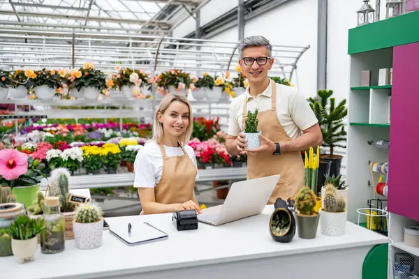 İki çiçekçi bir dükkânda çiçek ve bitki satışını sunuyorlar. Beyaz çalışanlar mutlu ve kameraya gülümsüyor..