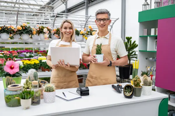 Çiçekçi ve asistan tezgahın önünde duruyor, çiçeklerle ve bitkilerle dolu, çiçekçilerinde müşterilerine hizmet etmeye hazır..