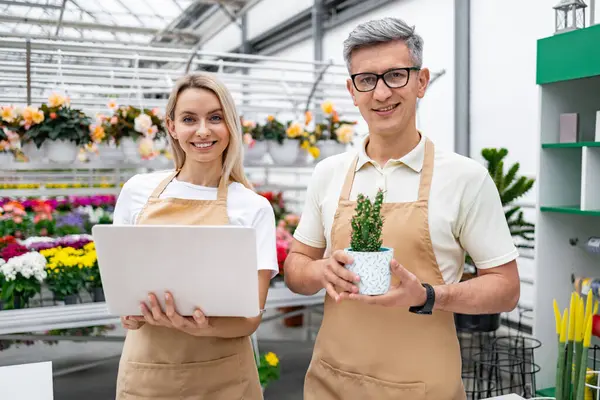 Bir çiçekçi ve bir meslektaşı, dükkanlarında dizüstü bilgisayar ve küçük bir saksı bitkisiyle poz verirken gülümsüyorlar..