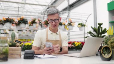 Çiçekçi dükkanındaki beyaz bir adam kazandıklarını sayıyor. Etrafı renkli çiçekler ve satılık bitkilerle çevrili. Tezgahta..
