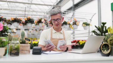 Çiçekçi dükkanındaki beyaz bir adam tezgahta nakit satışlarını memnuniyetle sayar..