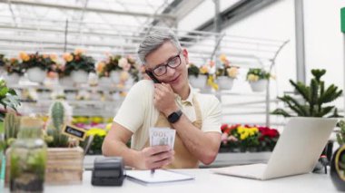 Telefondaki beyaz bir çiçekçi çiçekçi çiçekçi dükkanında bir dizüstü bilgisayar ve arka planda bitkilerle birlikte nakit sayıyor..