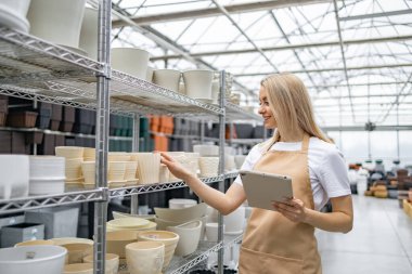 Önlüklü bir kadın dükkandaki saksılara bakar. Elinde bir tablet var..