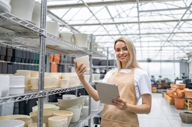Bir bitki dükkanında elinde saksı ve tablet tutan gülümseyen beyaz bir kadın. Raflar çeşitli saksılarla dolu..