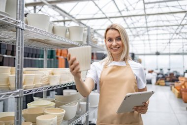 Önlüklü sarışın, beyaz bir kadın bir dükkandan saksı seçiyor. Elinde bir tablet var, izleyiciye gülümsüyor, çeşitli bitki konteynırlarını gösteriyor..