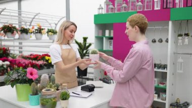 Çiçekçi dükkanı sahnesinde gülümseyen bir kadın ve kaktüslü bir kasiyerin yer aldığı bir satış işlemi yer alıyor..