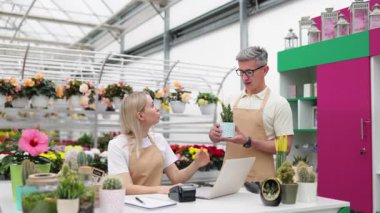 İki çiçekçi bir dükkânda çiçek ve bitki satışını sunuyorlar. Beyaz çalışanlar mutlu ve kameraya gülümsüyor..
