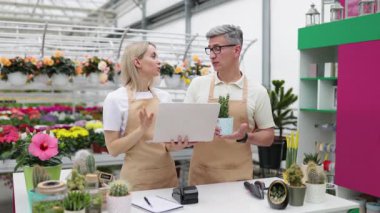 Bir çiçekçi ve bir müşteri mağazadaki satıştayken dizüstü bilgisayarı kontrol ediyorlar. Kadın dizüstü bilgisayarı tutuyor, ve adam saksı bitkisini tutuyor..