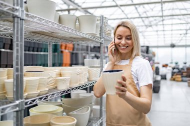 Beyaz bir kadın, çiçekçi dükkanında telefonuyla konuşurken saksıyı inceliyor. Arkadaki raflarda başka saksılar da var..