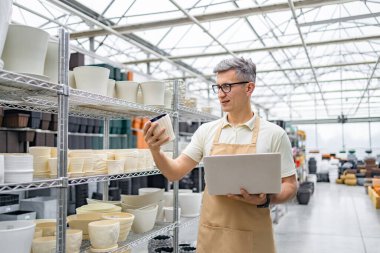 Kafkasyalı bir adam elinde dizüstü bilgisayarla raflarda birden fazla saksı bulunan bir dükkanda saksıyı inceliyor..