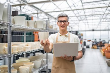 Çiçek saksısı ve dizüstü bilgisayar tutan bir dükkan sahibi, çeşitli seramik saksılarla çevrili, bir fabrikada. Adam gülümsüyor ve önlük giyiyor..