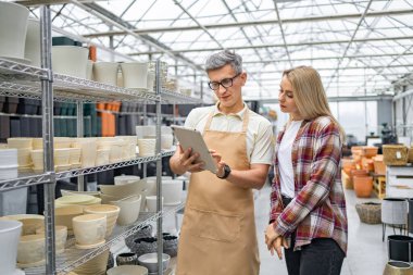 Bir tezgahtar müşterinin saksı seçmesine yardım eder. Çift bir tablete bakıyor. Bitki saksıları görünür durumda..