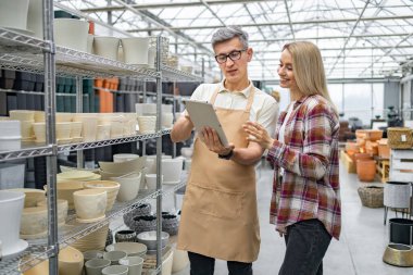Bir müşteri, bir çiçekçi dükkanındaki saksılar hakkında, bir tabletin yardımıyla, parlak bir ticari ortamda, bir mağaza asistanına danışmanlık yapar..