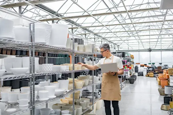 Beyaz bir adam elinde dizüstü bilgisayar ve önlükle dükkandaki saksıları inceliyor. Sıralar dolusu saksı rafları dolduruyor..