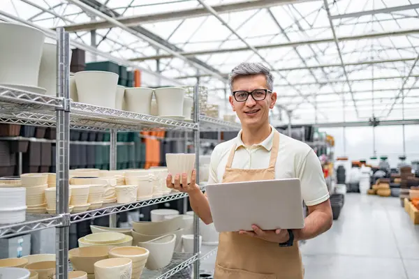 Gözlüklü beyaz bir adam saksı dolu bir dükkanda elinde bir saksı ve dizüstü bilgisayar tutuyor..