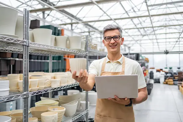 Çiçekçide gülümseyen bir adam elinde bir saksı ve dizüstü bilgisayarla arkasında başka saksılar tutuyor..