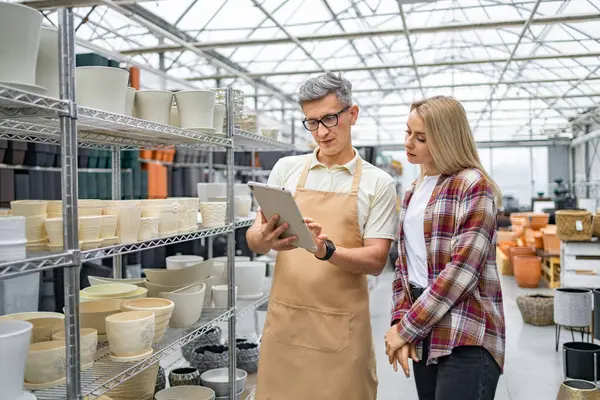 Bir tezgahtar müşterinin saksı seçmesine yardım eder. Çift bir tablete bakıyor. Bitki saksıları görünür durumda..