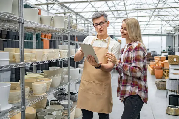 Bir müşteri, bir çiçekçi dükkanındaki saksılar hakkında, bir tabletin yardımıyla, parlak bir ticari ortamda, bir mağaza asistanına danışmanlık yapar..