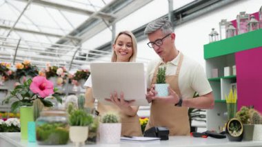 İki çiçekçi, bir erkek ve bir kadın, bir fabrikada dizüstü bilgisayara bakıyorlar, masada saksıdaki küçük bir bitkiden bahsediyorlar..