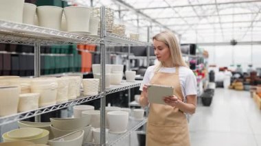 Önlüklü bir kadın dükkandaki saksılara bakar. Elinde bir tablet var..