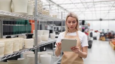 Bir tezgahtar saksıları tabletle inceliyor. Mağazada çeşitli saksılar var. Dükkanda cam tavan var..