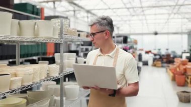 Kafkasyalı bir adam, elinde envanter için dizüstü bilgisayarı olan bir çiçekçide saksıyı inceliyor. Dükkan çeşitli tencerelerle dolu raflarla düzenlenir..