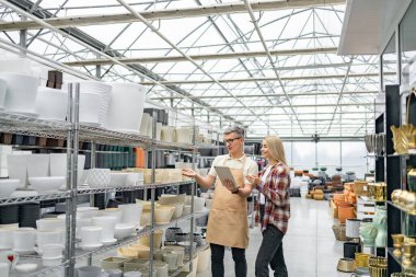 Bir erkek ve kadın bir dükkanda çiçek saksılarını karıştırarak seçeneklerini tartışıyor. Dükkan çok çeşitli tencere satar..