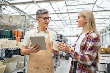 Bir tezgahtar, bir saksı seçmede müşteriye yardımcı olur, bir tablet için yardım önerir. İç mekanda şık tencereler sergileniyor..