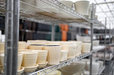 Bir fabrikadaki raflar farklı boyutlarda ve tasarımlarda çeşitli saksılar sergiliyor, hepsi satışa hazır..