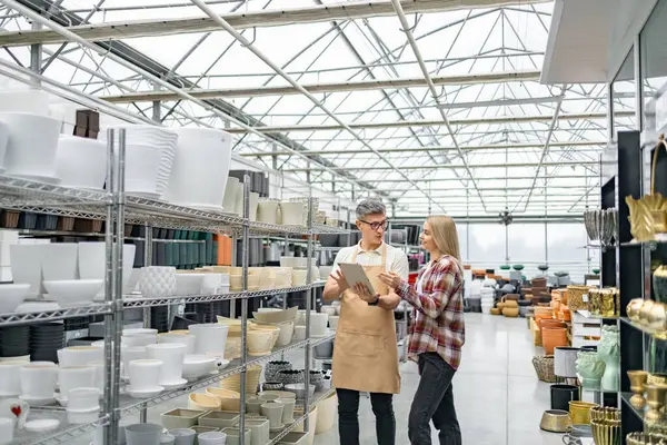 Bir tezgahtar bir mağazada bir müşteriyle saksı hakkında konuşur. Raflar çeşitli, şık saksılarla dolu..