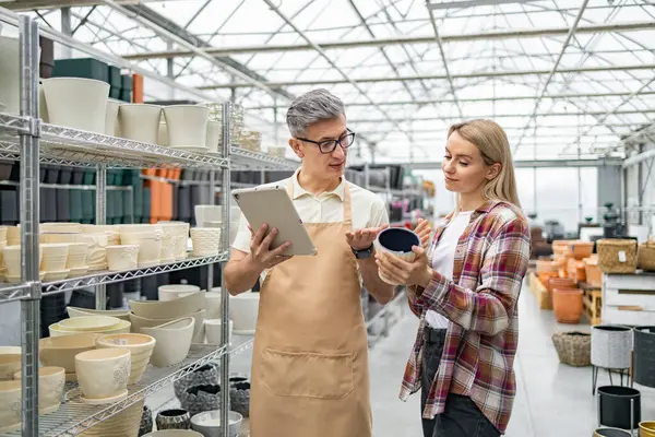 Bir tezgahtar, müşteriye çiçekçiden çiçek saksısı seçmesinde yardımcı olur ve mevcut seçenekleri tartışır..