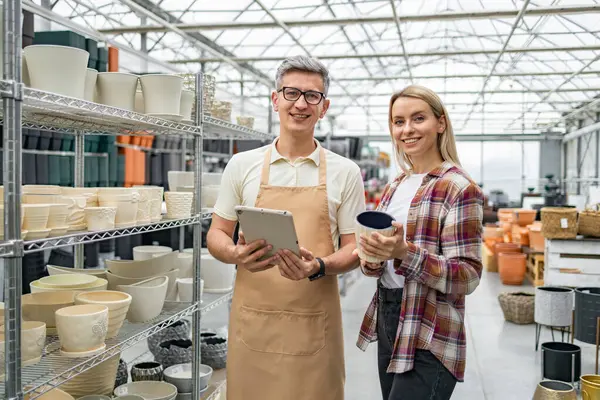 Bir mağaza çalışanı, çeşitli bitki konteynırlarıyla çevrili bir bahçe merkezindeki saksıları seçen müşteriye yardım eder..