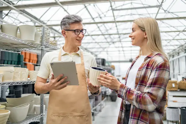 Bir tezgahtar, bir saksı seçmede müşteriye yardımcı olur, bir tablet için yardım önerir. İç mekanda şık tencereler sergileniyor..