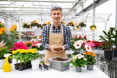 Serada gülümseyen bir adam canlı çiçekler, saksılar ve bahçe aletleriyle çevrili toprağı sergiler..