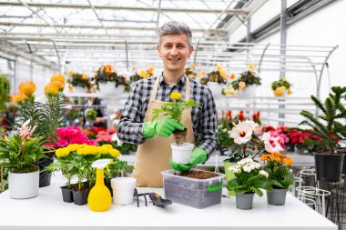 Bir serada gülümseyen bir adam, çeşitli çiçek ve bahçe aletleriyle çevrili bir saksı çiçeği tutar..