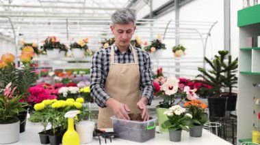Serada gülümseyen bir adam canlı çiçekler, saksılar ve bahçe aletleriyle çevrili toprağı sergiler..
