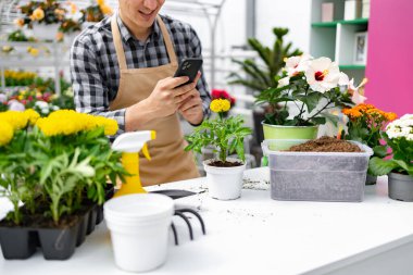 Bir çiçekçi, çeşitli bitki ve bahçe aletleriyle çevriliyken telefonda bir çiçeğin fotoğrafını çekiyor..