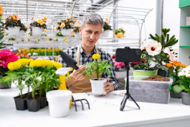 Bir adam serada video çekerken bir bitki gösteriyor. Çiçekler ve bahçe aletleri beyaz bir masada..
