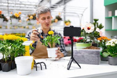 Adamın biri akıllı telefonuyla bahçe işleriyle ilgili bir video kaydediyor. Bitkiler ve bahçe aletleri masanın üzerinde..