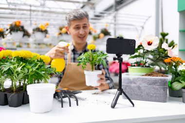 Bir bahçıvan, bir seradaki bitkilerle ilgili bir video kaydediyor. Sahnede çeşitli çiçek ve bahçe aletleri yer alıyor.